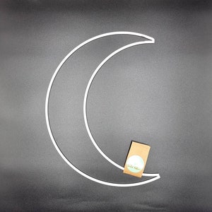 Könnte beinhalten: Eine weiße Metall-Halbmondform mit einem kleinen braunen Papieretikett, das an der Unterseite befestigt ist. Das Etikett hat einen weißen Kreis mit dem Text "Sweet William" in Grün.