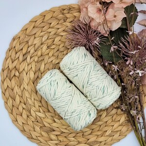 Puede incluir: Dos bobinas de hilo de macramé blanquecino con motas verdes descansan sobre un salvamanteles tejido. Flores y follaje rosas secos están dispuestos alrededor del hilo. El hilo es un suministro artesanal para proyectos de macramé.