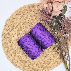 Puede incluir: Dos madejas de hilo morado vibrante descansan sobre un salvamanteles circular tejido. Flores secas en tonos rosas y morados están dispuestas alrededor del hilo. El fondo es una superficie blanca lisa.