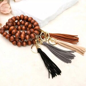 May include: A collection of wooden bead bracelets and keychains with tassel accents. The keychains feature gold-colored hardware and tassels in black, gray, tan, and brown. The wooden beads are a warm brown color.