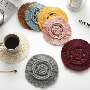 May include: A collection of handmade macrame coasters in various colors, including beige, brown, gray, yellow, pink, and burgundy. The coasters have a circular design with a central spiral and fringed edges. A coffee cup and a gold spoon are also visible.
