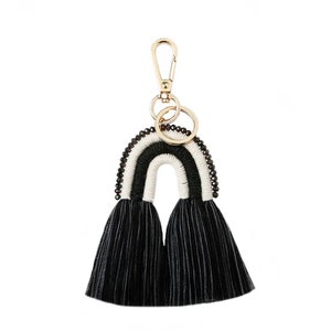 May include: A black and white rainbow-shaped keychain with a gold-colored clasp and key ring. The rainbow design features a white center, black stripes, and black beaded accents. The bottom of the keychain has two black tassels.