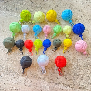 May include: A collection of colorful monkey fist keychains in various shades, including green, yellow, blue, pink, and black. Each keychain features a tightly knotted ball design with a metal clasp for easy attachment. The keychains are arranged on a textured surface.