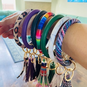 May include: A collection of key rings with various designs. The key rings feature a variety of patterns, including leopard print, marble, and stripes. Each ring has a gold-colored clasp and a matching tassel.