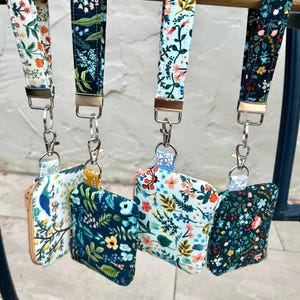 May include: Four floral print lanyards with matching small zipped pouches. The lanyards and pouches feature various colourful floral patterns on white, navy, and light blue backgrounds. Each pouch is attached to the lanyard with a silver-coloured metal clasp.