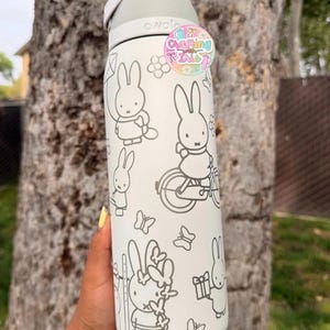 May include: A white Owala water bottle featuring a gray line art pattern of Miffy the rabbit in various activities, including riding a bicycle and holding a gift. A circular sticker with the text Crafting with Isle is attached to the top.