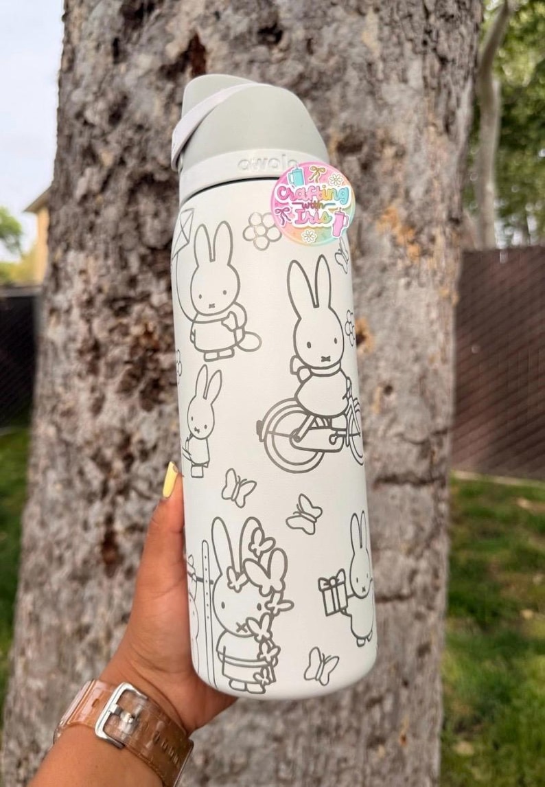 May include: A white Owala water bottle featuring a gray line art pattern of Miffy the rabbit in various activities, including riding a bicycle and holding a gift. A circular sticker with the text Crafting with Isle is attached to the top.