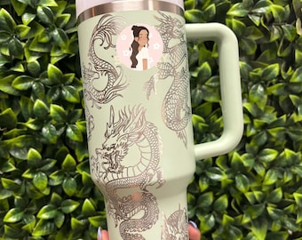 May include: A mint green tumbler with a handle, straw, and lid. The tumbler is decorated with silver dragon designs and a small sticker of a woman. The background is a wall of green leaves.