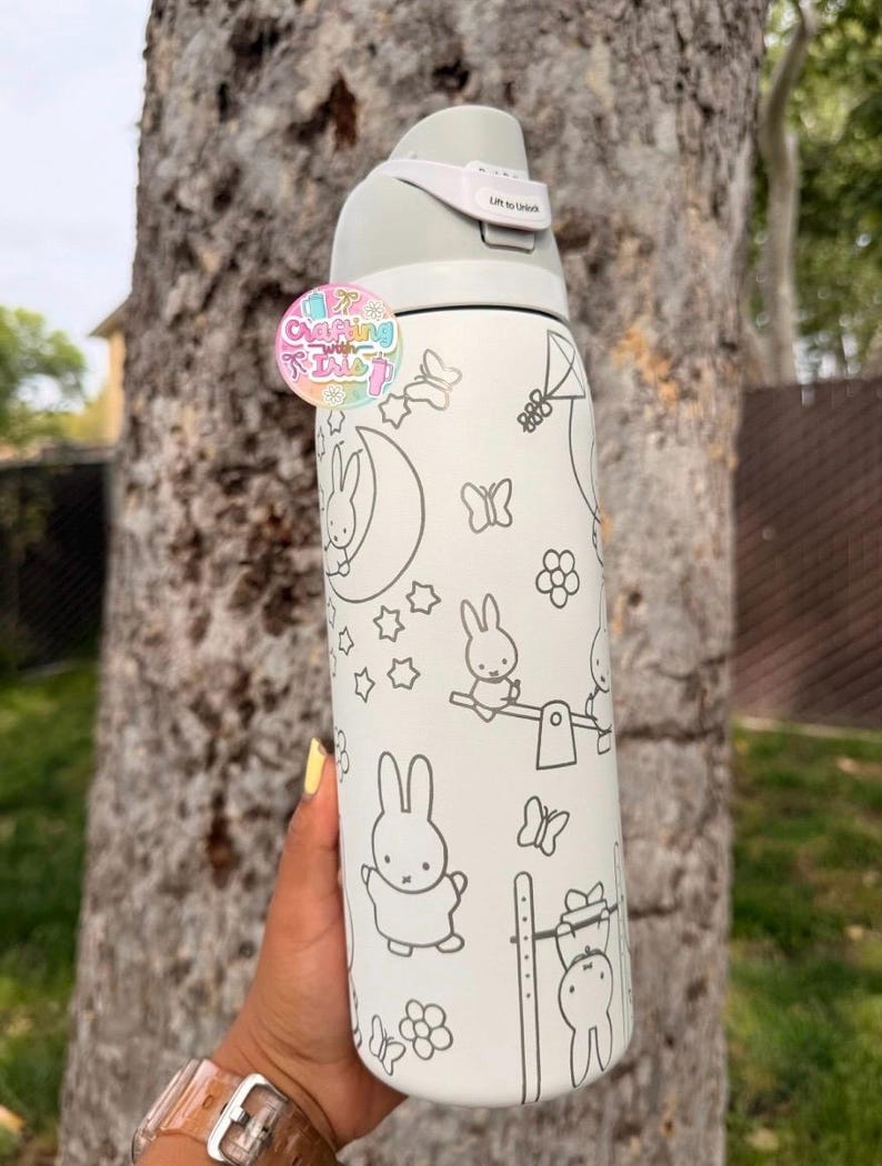 May include: A white stainless steel water bottle featuring a gray line art pattern of rabbits, butterflies, stars, and flowers. The bottle has a gray lid with a locking mechanism labeled Lift to Unlock and a circular logo sticker attached.