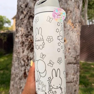 May include: A white Owala water bottle featuring black line art of Miffy characters, flowers, butterflies, and balloons. The bottle has a light gray lid and a circular sticker near the top that reads Crafting with Iris in colorful letters.