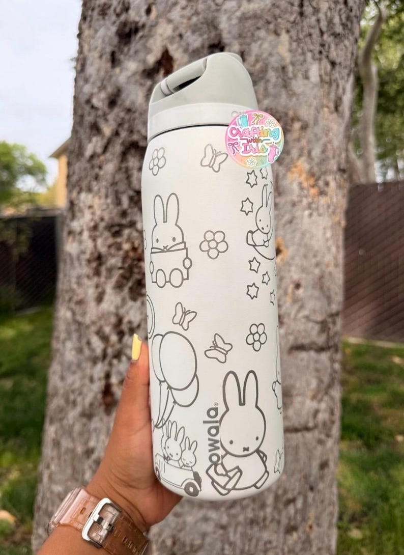 May include: A white Owala water bottle featuring black line art of Miffy characters, flowers, butterflies, and balloons. The bottle has a light gray lid and a circular sticker near the top that reads Crafting with Iris in colorful letters.