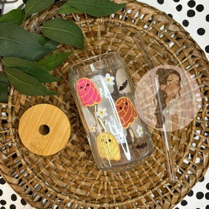May include: A clear glass tumbler with a bamboo lid and a glass straw. The tumbler has a design of cartoon ghosts with different colored pastry fillings. The ghosts are surrounded by white flowers and bats.