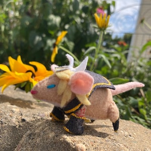 May include: A handmade felt creature with a tan body, pink ears, and a long pink tail. It has black and yellow accents on its armor and boots. The creature is posed on a rock with yellow flowers in the background.