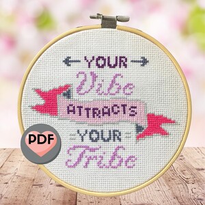 May include: A cross-stitch embroidery hoop with the text "YOUR Vibe ATTRACTS YOUR Tribe" in purple thread. The design includes pink arrow accents and a PDF icon. The hoop is set against a blurred floral background.