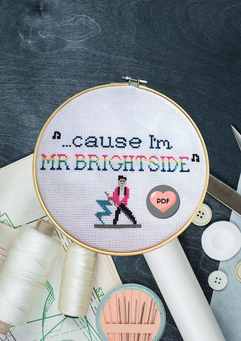 May include: Cross stitch pattern featuring a pixelated illustration of a person in a pink jacket and black pants holding a microphone. The text "...cause I'm MR. BRIGHTSIDE" is stitched in rainbow colors. A pink heart with the word "PDF" is in the lower right corner.