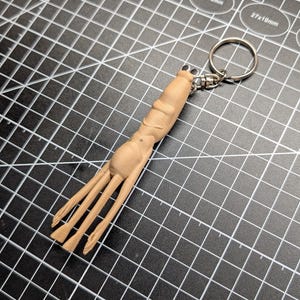 May include: A light brown wooden keychain featuring a stylized, abstract design. The keychain has a ring for attaching keys and a carved, organic shape with three prongs extending from the base. The item is set against a black grid.