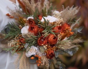 May include: A bridal bouquet with a mix of white and orange flowers, dried grasses, and greenery. The bouquet is held by a person wearing a white dress.