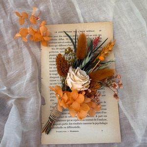 May include: A bouquet of dried flowers in shades of orange, brown, and cream, tied with twine. The arrangement sits on a vintage book page with French text. The bouquet includes a variety of textures and shapes, creating a rustic aesthetic.