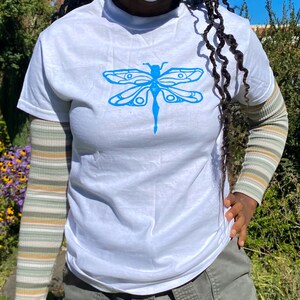 Puede incluir: Camiseta blanca con un estampado gr&aacute;fico de lib&eacute;lula azul.