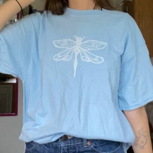 Puede incluir: Una camiseta azul claro con un dise&ntilde;o gr&aacute;fico de lib&eacute;lula blanca.