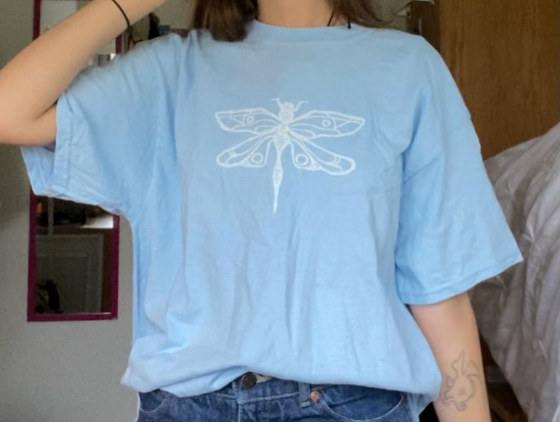 Puede incluir: Una camiseta azul claro con un dise&ntilde;o gr&aacute;fico de lib&eacute;lula blanca.