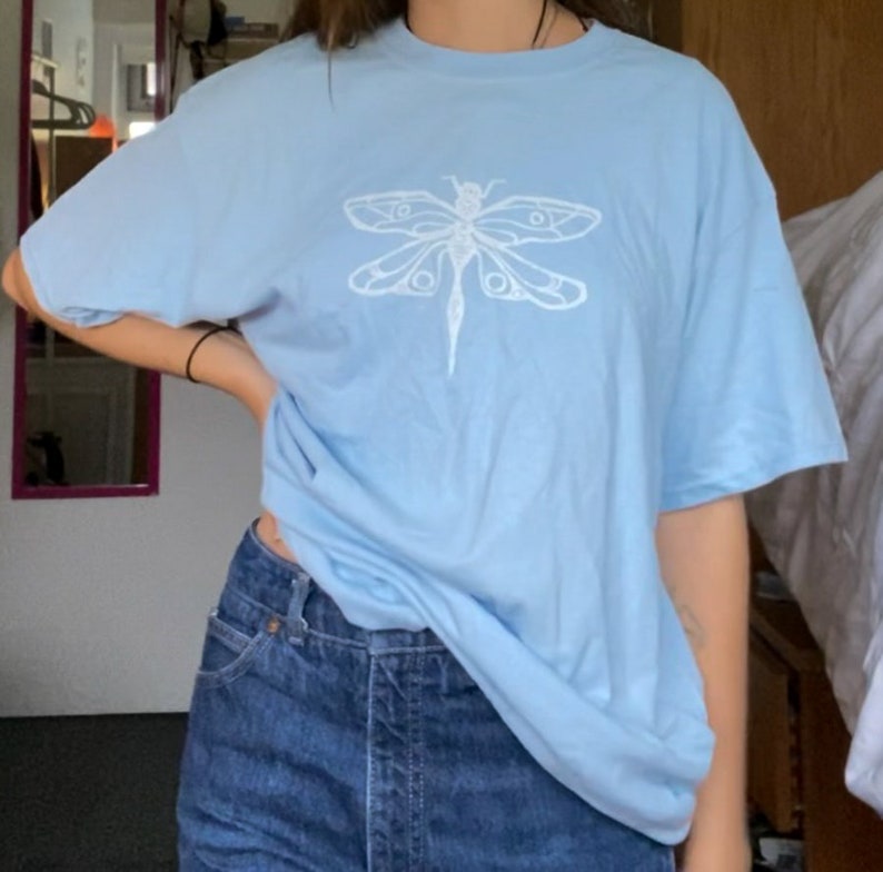 Puede incluir: Una camiseta azul claro con un dise&ntilde;o gr&aacute;fico de lib&eacute;lula blanca.