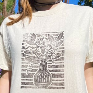 May include: A white t-shirt with a black and white print of a vase with flowers. The print is in a block style and has a striped background.