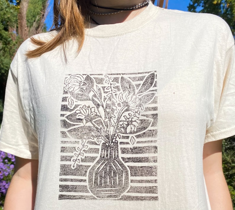 May include: A white t-shirt with a black and white print of a vase with flowers. The print is in a block style and has a striped background.