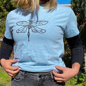 Puede incluir: Una camiseta azul claro con un gr&aacute;fico de lib&eacute;lula negro impreso en la parte delantera.