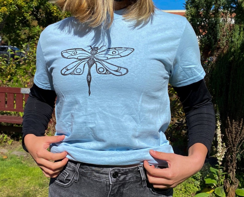Puede incluir: Una camiseta azul claro con un gr&aacute;fico de lib&eacute;lula negro impreso en la parte delantera.