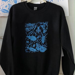 May include: Black sweatshirt with a blue insect print. The design features various insects, including butterflies, bees, and beetles. The sweatshirt is hanging on a hanger against a white wall with blue art prints.
