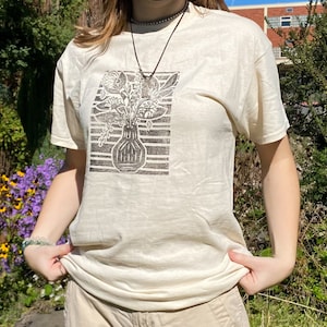 May include: A white t-shirt with a black and white print of a vase with flowers. The print is in a block style.