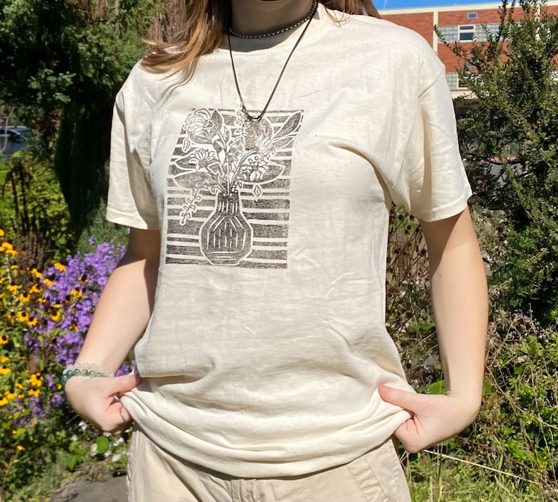 May include: A white t-shirt with a black and white print of a vase with flowers. The print is in a block style.