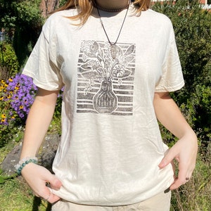 May include: A white t-shirt with a black and white print of a vase with flowers. The print is a block print style.