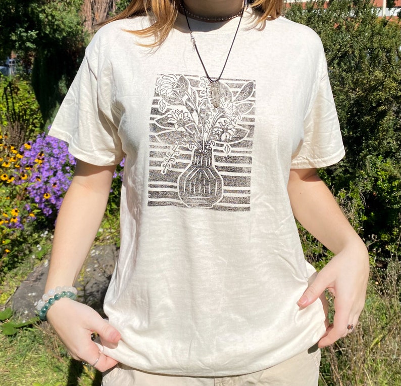 May include: A white t-shirt with a black and white print of a vase with flowers. The print is a block print style.