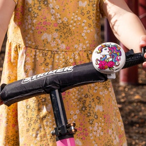May include: A child riding a black and pink Strider bike with a unicorn bell. The bell is white with a colorful unicorn design and pink flowers.
