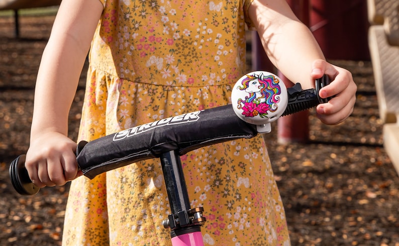 May include: A child riding a black and pink Strider bike with a unicorn bell. The bell is white with a colorful unicorn design and pink flowers.