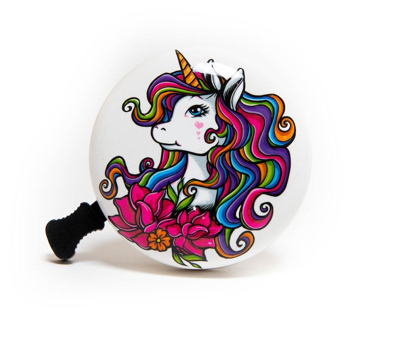 May include: White bicycle bell with a colorful unicorn design. The unicorn has a rainbow mane and tail, and is surrounded by pink flowers.