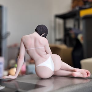 May include: A figurine of a person in a white bikini, lying down with their back to the viewer. The figure has dark hair and is posed on a reflective surface.