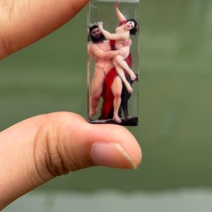 Peut inclure: Pendentif rectangulaire en résine représentant une sculpture miniature d'un homme et d'une femme dans une étreinte passionnée. Les personnages sont nus, la femme portant une robe rouge. Le pendentif est tenu entre les doigts sur un fond vert flou.