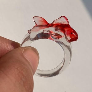 Peut inclure: Une bague en résine transparente ornée d'un poisson rouge. Le poisson rouge a des nageoires détaillées et un petit œil. La bague est tenue par une main sur fond blanc. Un bijou unique.