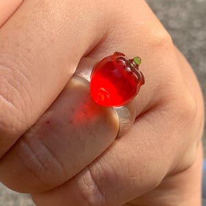 Peut inclure: Une bague transparente ornée d'un charm en forme de fraise rouge avec une tige verte. La bague est tenue dans une main. Un bijou unique.