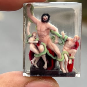Peut inclure: Petite sculpture dans de la résine transparente. Elle représente une figure musclée avec une barbe, entourée de deux petites figures et de serpents verts. Les personnages sont sur une base sombre, avec des détails rouges et blancs.