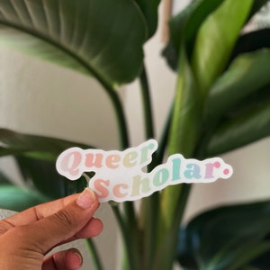 May include: A sticker with the words "Queer Scholar" in pastel colors. The sticker is held in front of a green plant. The text is in a playful, rounded font, with each word in a different pastel hue.