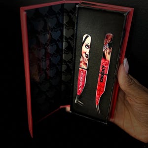May include: A red and black box containing two pairs of eyelash tweezers. The tweezers are decorated with a red and black pattern and the words "Chucky" and "Eat Your Heart Out".