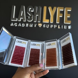 Könnte beinhalten: Eine Hand hält eine transparente, mehrteilige Hülle mit vier verschiedenen Farbtönen von Wimpernverlängerungen. Die Hüllen sind mit "Lash Lyfe" beschriftet und enthalten verschiedene Brauntöne und rote Wimpern. Der Hintergrund zeigt das Logo "Lash Lyfe Academy Supplies".