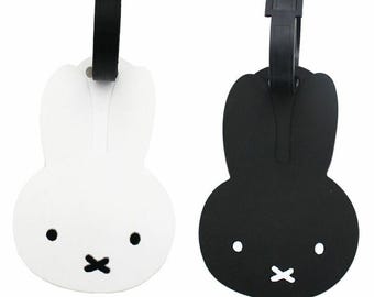 May include: Two luggage tags shaped like a bunny, one white and one black. Each tag has a black strap and buckle for attachment. The bunny face features two black dots for eyes and a black 'x' for a mouth. The word 'miffy' is printed in orange.