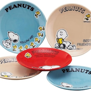May include: A collection of ceramic plates featuring Peanuts characters. The plates are in various colors, including blue, tan, and red. Each plate has the word "PEANUTS" and images of Snoopy and Woodstock. Some plates have text that says "FOLLOW ME!" and "BEST FRIENDS."