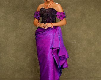 Könnte beinhalten: Eine Frau in einem traditionellen nigerianischen Outfit. Das Ensemble umfasst ein lila-schwarzes schulterfreies Oberteil, einen passenden Wickelrock und ein Kopftuch. Das Outfit ist mit orangefarbenem Perlschmuck und lila floralen Akzenten versehen.