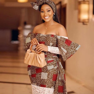 May include: A woman wearing a patterned off-the-shoulder dress with a matching head wrap. The dress features a brown, red, and white geometric design. She is holding a tan handbag and wearing a gold necklace and earrings.